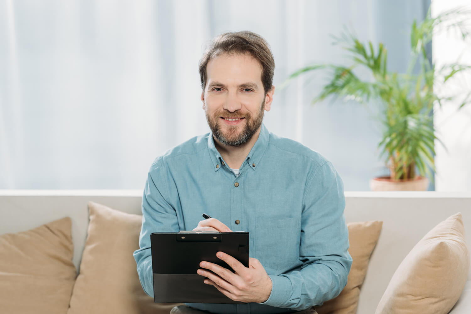 A man with a beard, wearing a light blue shirt, is sitting on a couch holding a clipboard and pen. He is smiling, and there is a potted plant in the background.