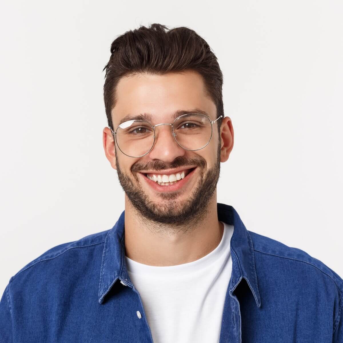 A smiling man with short brown hair and a beard wears clear-framed glasses and a denim shirt over a white t-shirt. The background is plain and white.