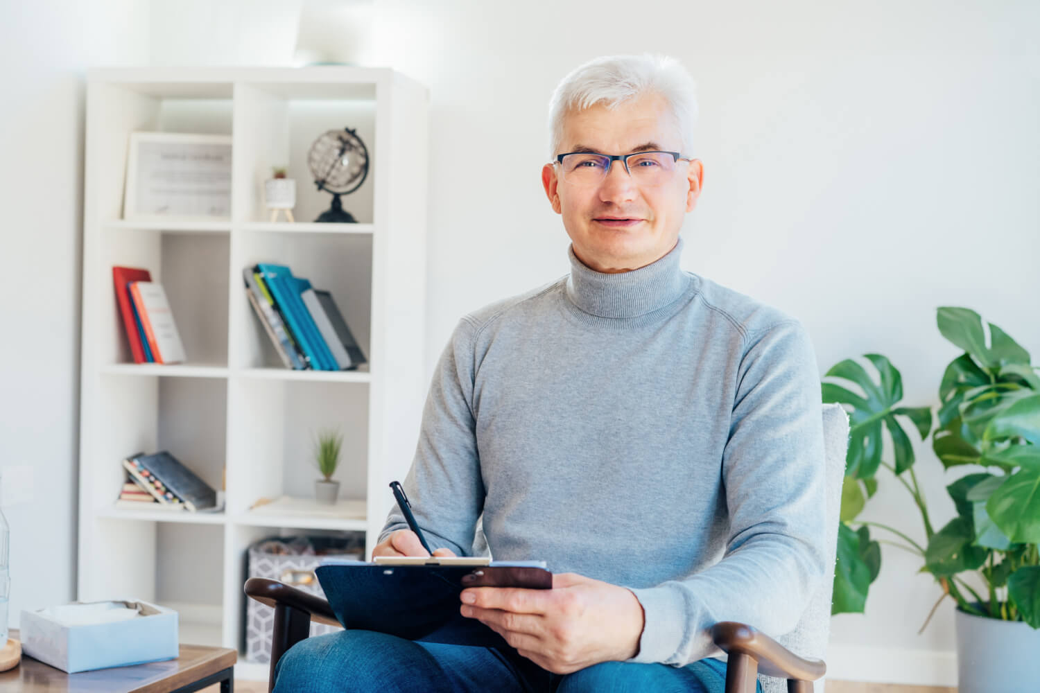 Man with glasses and gray hair sits smiling in a modern office. Hes wearing a gray turtleneck and holding a clipboard with a pen. In the background, there is a white bookshelf with books and decor, and a potted plant.