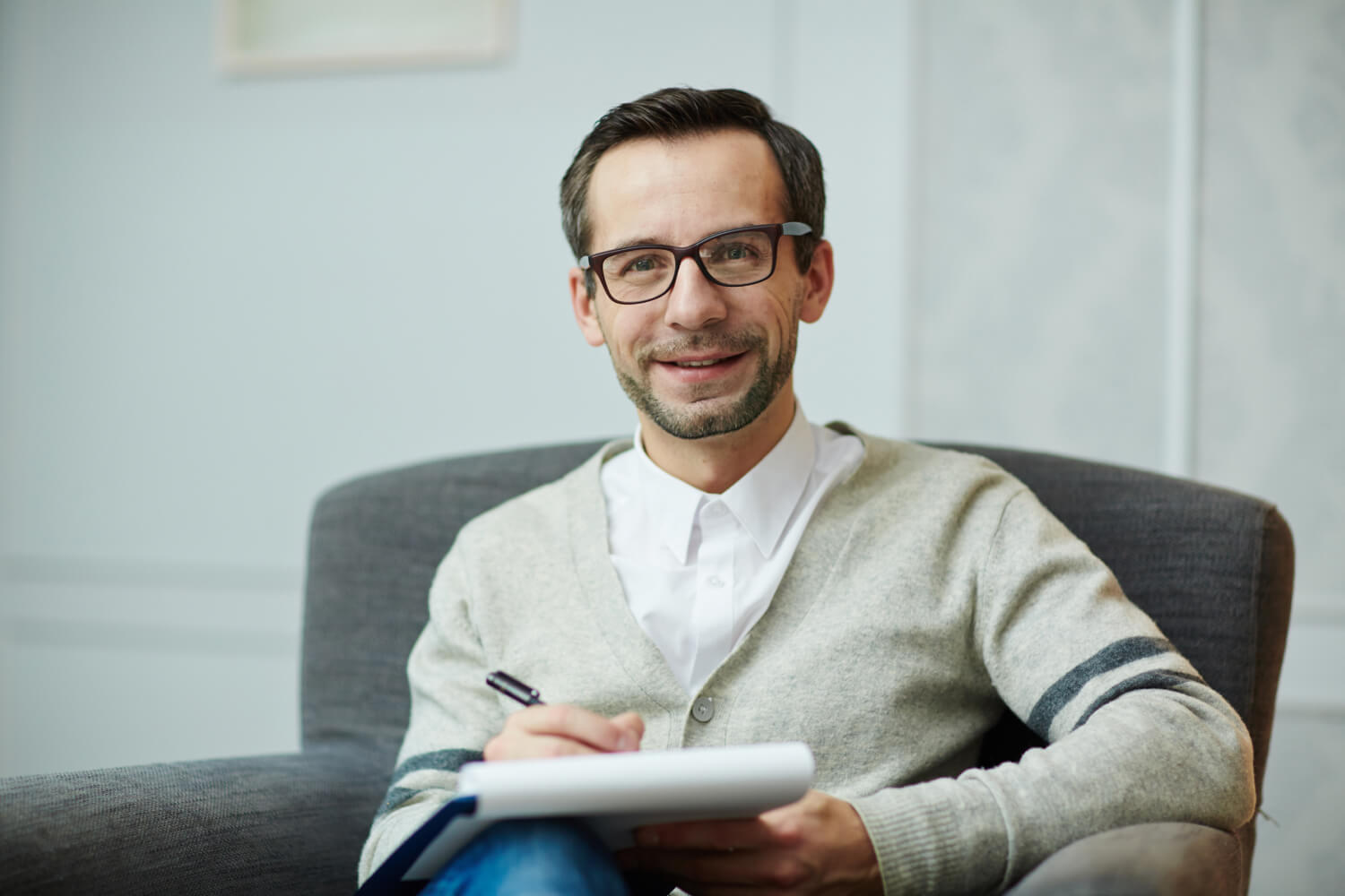 Smiling man with glasses, wearing a light sweater, sits in a chair holding a notebook and pen. He appears relaxed in a softly lit room with a neutral background.