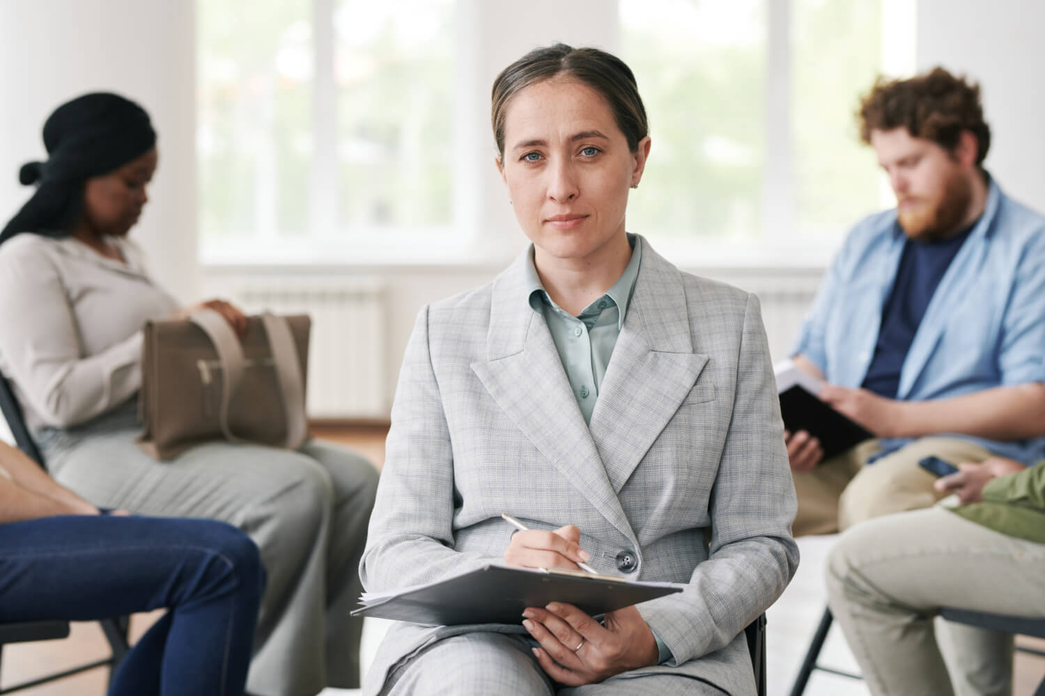 A person in a gray suit sits in a bright room, holding a clipboard and looking at the camera. Three others in casual clothes sit nearby, engaged in different activities, such as reading or holding a bag. The room has large windows in the background.